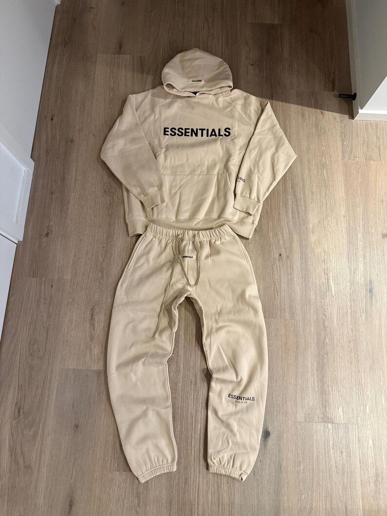 May include: A beige hoodie and matching sweatpants set. The word "ESSENTIALS" is printed in black on the front of the hoodie and on the left leg of the sweatpants. The hoodie has a drawstring hood and a front pocket.