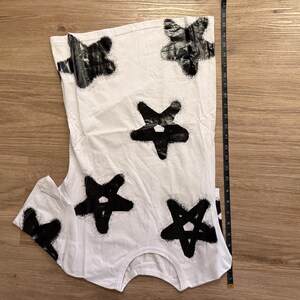 May include: White t-shirt with a black star pattern. The stars are irregularly shaped and appear to be spray-painted. A measuring tape is placed next to the shirt, indicating its length.