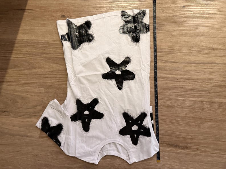May include: White t-shirt with a black star pattern. The stars are irregularly shaped and appear to be spray-painted. A measuring tape is placed next to the shirt, indicating its length.