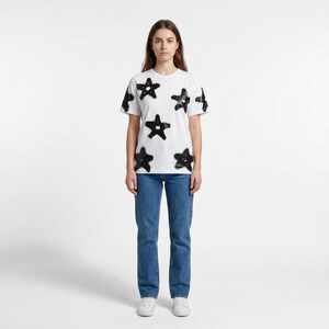 May include: A white t-shirt with black star designs is paired with blue jeans. The short-sleeved shirt features multiple black star shapes across the front and sleeves. The model is wearing white sneakers.