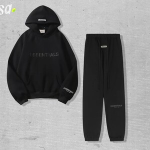 May include: Black hoodie and sweatpants set. The hoodie features the word "ESSENTIALS" across the chest and the brand name on the sleeve. The sweatpants have a drawstring waist and the brand name on the leg. The set is made of a soft material.