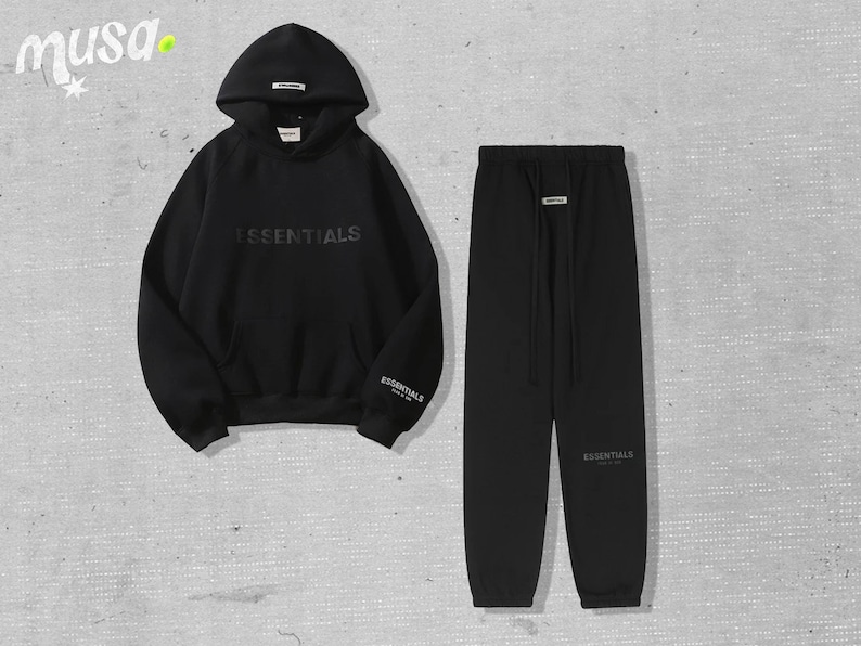 May include: Black hoodie and sweatpants set. The hoodie features the word "ESSENTIALS" across the chest and the brand name on the sleeve. The sweatpants have a drawstring waist and the brand name on the leg. The set is made of a soft material.
