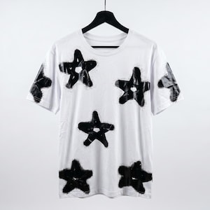 May include: White t-shirt with a crew neck and short sleeves, adorned with multiple black star designs. The stars have a spray-painted effect, creating a unique, artistic look. The shirt is hanging on a black hanger.
