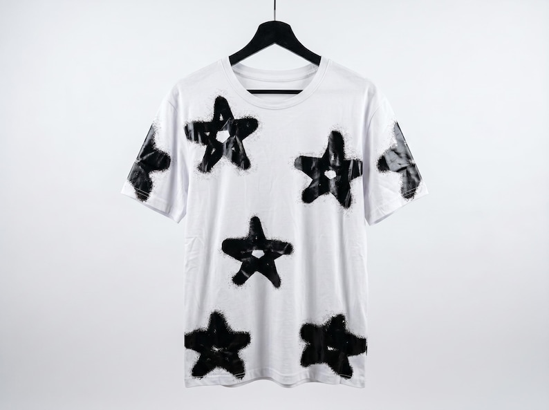 May include: White t-shirt with a crew neck and short sleeves, adorned with multiple black star designs. The stars have a spray-painted effect, creating a unique, artistic look. The shirt is hanging on a black hanger.