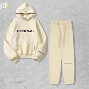 May include: A cream-colored hoodie and matching sweatpants set. The hoodie features the word "ESSENTIALS" in black block letters across the chest and the word "ESSENTIALS" on the left sleeve. The sweatpants have a drawstring waist and the word "ESSENTIALS" printed on the left leg.