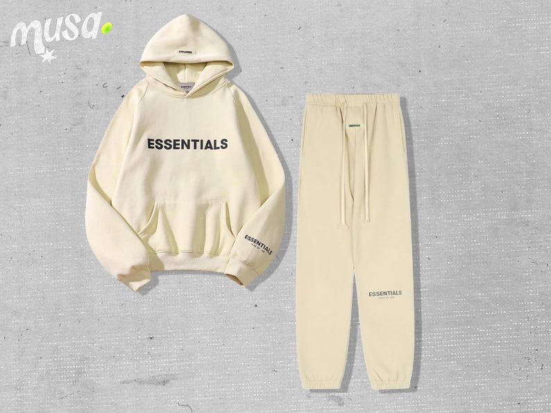 May include: A cream-colored hoodie and matching sweatpants set. The hoodie features the word "ESSENTIALS" in black block letters across the chest and the word "ESSENTIALS" on the left sleeve. The sweatpants have a drawstring waist and the word "ESSENTIALS" printed on the left leg.