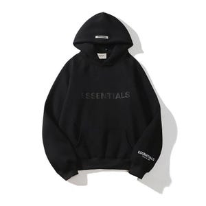 May include: Black hoodie with the word "ESSENTIALS" across the chest and on the left sleeve. The hoodie has a hood with a small tag at the top and a front pocket. The hoodie is made of a soft material.