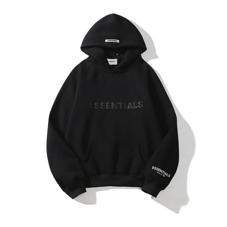 May include: Black hoodie with the word "ESSENTIALS" across the chest and on the left sleeve. The hoodie has a hood with a small tag at the top and a front pocket. The hoodie is made of a soft material.