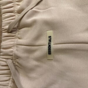May include: Close-up of beige sweatpants with a drawstring waistband. The pants have a small rectangular tag that reads "ESSENTIALS" in black text. The fabric appears soft and slightly textured.