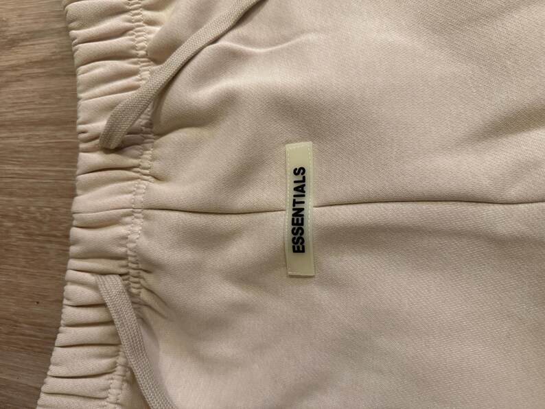 May include: Close-up of beige sweatpants with a drawstring waistband. The pants have a small rectangular tag that reads "ESSENTIALS" in black text. The fabric appears soft and slightly textured.