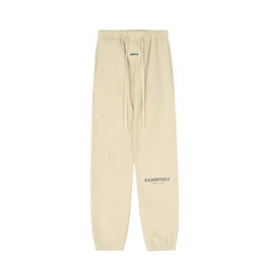 May include: Tan sweatpants with a drawstring waist and the word "ESSENTIALS" printed on the left leg. The pants are made of a soft material and have a relaxed fit. The pants are a neutral color.