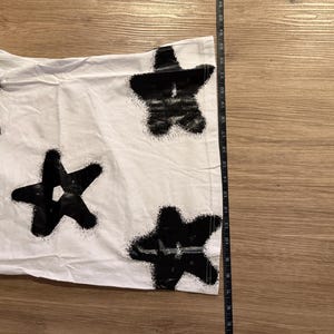 May include: White t-shirt with a black star pattern. The stars have a spray-painted effect. The shirt is laid flat on a wooden surface. A ruler is placed next to the shirt to measure its length.