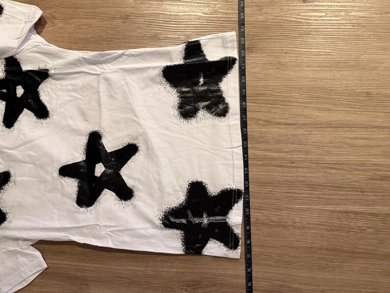 May include: White t-shirt with a black star pattern. The stars have a spray-painted effect. The shirt is laid flat on a wooden surface. A ruler is placed next to the shirt to measure its length.
