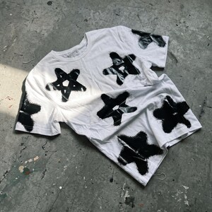 May include: White t-shirt with a black star pattern. The stars are irregularly shaped and appear to be spray-painted onto the fabric. The shirt is lying on a gray concrete surface, with a few paint splatters visible.