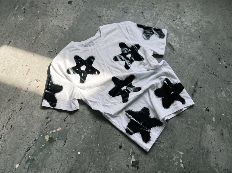 May include: White t-shirt with a black star pattern. The stars are irregularly shaped and appear to be spray-painted onto the fabric. The shirt is lying on a gray concrete surface, with a few paint splatters visible.