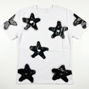 May include: White t-shirt with a crew neck and short sleeves. The shirt features a pattern of large, black, star-shaped designs. The stars have a textured, spray-painted appearance, creating a bold contrast against the white fabric.