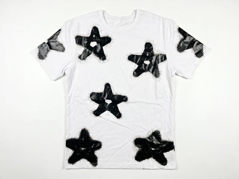 May include: White t-shirt with a crew neck and short sleeves. The shirt features a pattern of large, black, star-shaped designs. The stars have a textured, spray-painted appearance, creating a bold contrast against the white fabric.