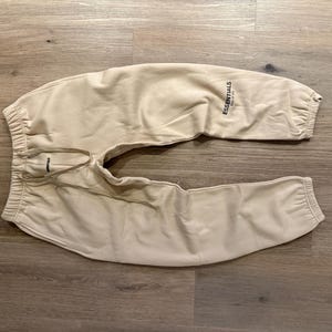 May include: Tan sweatpants with elastic cuffs and waistband, and a drawstring. The word "ESSENTIALS" is printed in black on the left leg. The pants are made of a soft material and are laid out on a wooden surface.