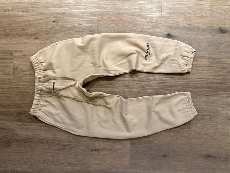 May include: Tan sweatpants with elastic cuffs and waistband, and a drawstring. The word "ESSENTIALS" is printed in black on the left leg. The pants are made of a soft material and are laid out on a wooden surface.