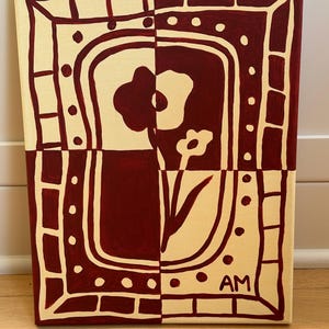 Puede incluir: Un cuadro rectangular sobre lienzo con un dise&ntilde;o dividido en granate oscuro y crema. La composici&oacute;n muestra un motivo floral estilizado dentro de un borde decorativo de puntos y rect&aacute;ngulos. Las letras AM est&aacute;n pintadas en la esquina inferior derecha.