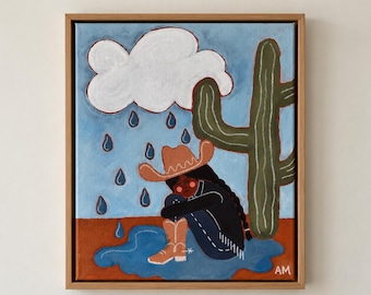 Puede incluir: Una pintura enmarcada sobre lienzo que representa una figura con sombrero de vaquero y botas marrones, sentada en un charco azul bajo una nube de lluvia blanca. Un cactus verde alto se encuentra a la derecha sobre un cielo azul claro y suelo marrón. Las iniciales AM están en la esquina.