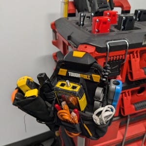May include: A black tool bag with a yellow and black ToughBuilt logo. The bag is hanging on a red toolbox and contains various tools, including a yellow cable tester, a black cable, and orange and white cables.