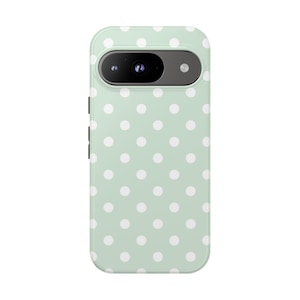 May include: A mint green phone case with a white polka dot pattern. The case has a camera cutout and button details on the side. The phone case is designed to fit a specific phone model.