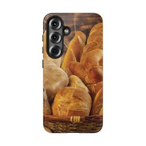 May include: A phone case featuring a basket filled with various types of bread. The bread includes loaves, rolls, and other baked goods in shades of brown and golden-yellow. The case has a dark brown border and a camera cutout.