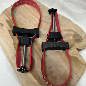 Op de afbeelding: Twee rode leren boeien met zwarte verstelbare onderdelen en zilveren spikes. De boeien liggen op een houten oppervlak. Eén is open, de andere gesloten. Waarschijnlijk voor bondage.