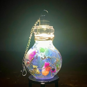 May include: A glowing, decorative glass bottle with a clear top and a silver chain. The bottle is filled with colorful objects and sits on a small black stand. The bottle is illuminated from within, creating a magical effect.