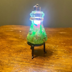 May include: A miniature, illuminated glass bottle with a green and pink interior, resting on a small black metal stand. The bottle is adorned with decorative elements and wrapped with twine. The light inside glows brightly.