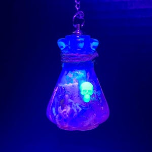 Pode incluir: Pingente azul brilhante em forma de frasco com uma caveira verde fluorescente. O pingente está pendurado em uma corrente prateada. O interior do frasco é adornado com desenhos roxos e azuis. Fundo azul escuro.