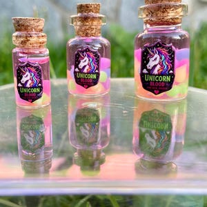 Puede incluir: Tres pequeñas botellas de vidrio con tapones de corcho, llenas de líquido rosa y capas coloridas. Cada botella tiene una etiqueta con un diseño de unicornio y las palabras "Unicorn Blood". Las botellas se reflejan en una superficie reflectante.