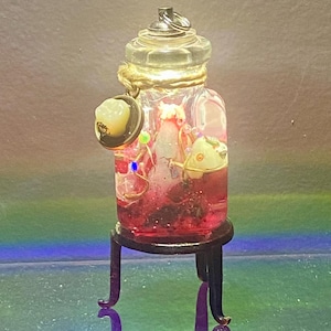 May include: A miniature glass jar with a clear lid, filled with a red liquid and small decorative elements. The jar is adorned with gold accents and sits on a small, black, ornate stand. The jar's contents include a small, white, tooth-shaped object.