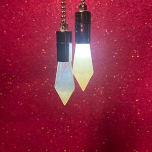 以下が含まれることがあります： ダイヤモンド型の光る照明器具が付いた、2つの装飾的なライトプルチェーン。 照明器具は白くキラキラしており、シルバーチェーンと黒いベースに取り付けられています。 背景はキラキラした濃い赤色です。