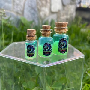 May include: Three small glass bottles with cork stoppers, filled with blue liquid and green orbs. Each bottle has a label that reads "Sea Serpent Blood" with a sea serpent illustration. The bottles are displayed on a clear, square stand.