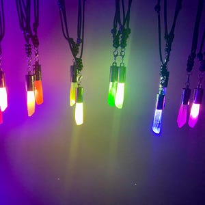 May include: Several glowing crystal pendant necklaces in various colors hang against a dark background. The crystals emit bright light in shades of red, orange, yellow, green, blue, and purple. Each necklace has a black cord.