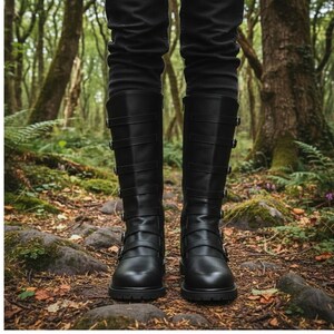 Puede incluir: Botas negras hasta la rodilla con m&uacute;ltiples correas y hebillas. Las botas tienen punta redonda y tac&oacute;n bajo. La persona lleva pantalones negros. Las botas est&aacute;n sobre un suelo forestal.