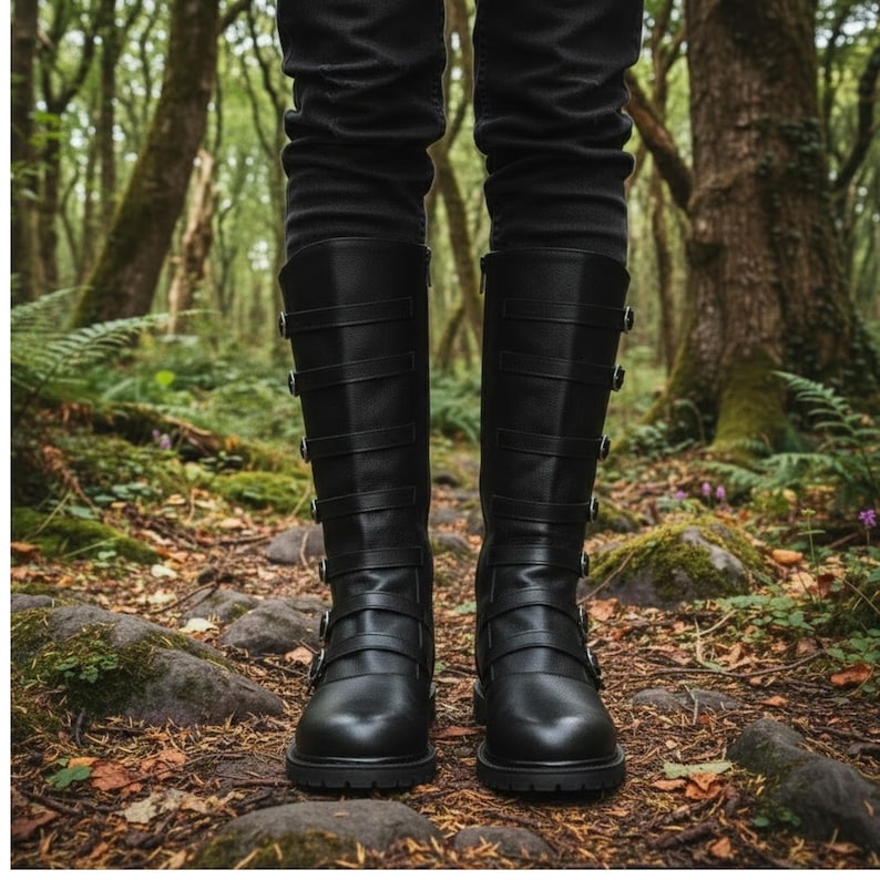 Puede incluir: Botas negras hasta la rodilla con m&uacute;ltiples correas y hebillas. Las botas tienen punta redonda y tac&oacute;n bajo. La persona lleva pantalones negros. Las botas est&aacute;n sobre un suelo forestal.