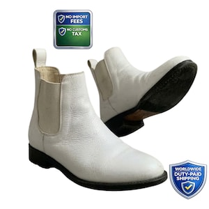 May include: A pair of white leather Chelsea boots with black soles. The boots have elastic side panels and pull tabs. The image also includes a badge that says "No Import Fees" and "No Customs Tax".