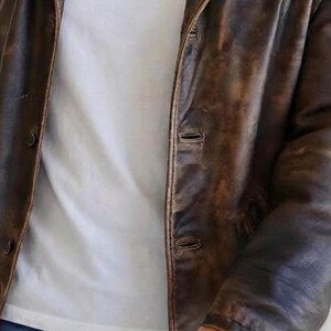 May include: A brown leather jacket with a distressed finish. The jacket has a button-up front and a classic collar. The jacket is worn over a white t-shirt. The leather appears to be worn and textured.