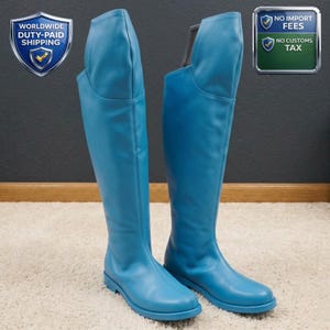 Puede incluir: Un par de botas altas de cuero azul claro. Las botas tienen un diseño hasta la rodilla con un acabado liso y mate. Las botas se muestran sobre una alfombra blanca. La imagen también incluye texto que dice "WORLDWIDE DUTY-PAID SHIPPING" y "NO IMPORT FEES NO CUSTOMS TAX".