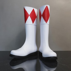 May include: Pair of white boots with red diamond patterns at the top. The boots have a black sole and a slight heel. The boots are made of a smooth, likely synthetic material, and are designed to reach the mid-calf.