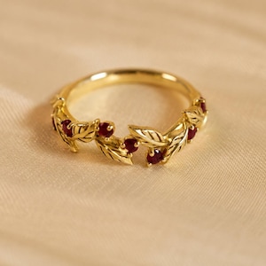 May include: A gold ring featuring a band designed with sculpted metal leaves and small, round, deep red rubies set between the foliage. The ring has a polished metallic finish and a continuous, vine-like structure that encircles the finger.