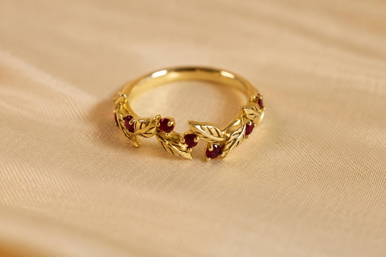 May include: A gold ring featuring a band designed with sculpted metal leaves and small, round, deep red rubies set between the foliage. The ring has a polished metallic finish and a continuous, vine-like structure that encircles the finger.