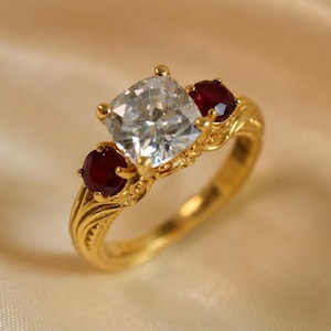 May include: A gold ring featuring a central cushion-cut clear gemstone flanked by two smaller round-cut deep red rubies. The gold band displays intricate, textured, carved detailing along the shank, providing a decorative finish to the metal.
