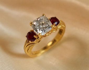 May include: A gold ring featuring a central cushion-cut clear gemstone flanked by two smaller round-cut deep red rubies. The gold band displays intricate, textured, carved detailing along the shank, providing a decorative finish to the metal.