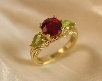 Könnte beinhalten: Ein Goldring mit einem großen, runden, tiefroten Mittelstein, flankiert von zwei kleineren, birnenförmigen, hellgrünen Steinen. Das Goldband zeigt komplizierte, erhabene Schnörkelmuster an den Seiten, die zu den Krappenfassungen führen.