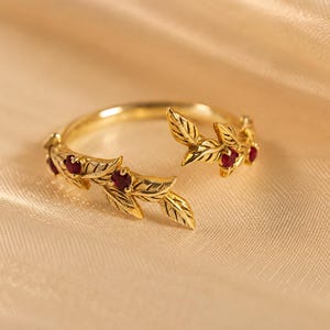 May include: A gold open-style ring featuring a band shaped like two leafy branches meeting in the center. The ring is adorned with small, round, deep red gemstones set within the gold leaves, creating a detailed botanical design.