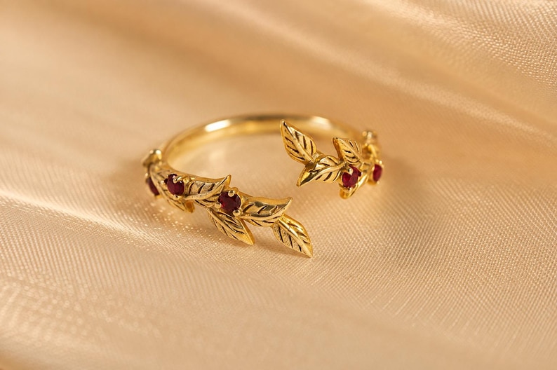 May include: A gold open-style ring featuring a band shaped like two leafy branches meeting in the center. The ring is adorned with small, round, deep red gemstones set within the gold leaves, creating a detailed botanical design.